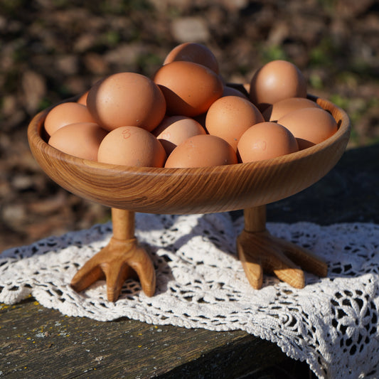 A wooden pedestal bowl filled with brown eggs, set on a white lace doily on a rustic wooden surface outdoors.